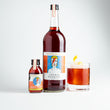 Load image into Gallery viewer, The small Aperol Negroni bottle is in the picture, alongside a large 750ml bottle. It is the same shape and similar label, but the bottle is a lot larger. Next to it is a glass of Aperol Negroni. It is a deep orange colour and is in a short tumbler glass with a large ice cube, and a lemon twist sitting on top of the ice cube. The background is white.