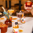 Load image into Gallery viewer, The Aperol Negroni is shown here in a glass being drunk by someone out of shot. There is a Lychee Martini in a nice nic and Nora glass on the table in what could be a house or a bar. There are other drinks and a vase of dried flowers blurred out on the table, and what looks like a bowl of almonds.