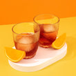 Load image into Gallery viewer, Two Aperol Negronis are in shot, in small short glasses with a ball of ice in each glass. They are on a short white stand, with two orange slices next to each glass. It's quite an arty shot, with the white stand being on a lemon yellow table, and the background is a nice orange colour. It's a bright, colourful fun picture. The cocktails look nicely chilled.