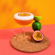 Load image into Gallery viewer, A nice bright picture of a passion fruit martini by Mindful Mixology. It is on a brown cork coaster, on a salmon pink table, with a deep orange background. The cocktail is in a coupe and is orange with a big frothy layer of foam on top that is white. A passionfruit half is floating in the drink. Next to the glass are 1.5 limes and half a passionfruit.