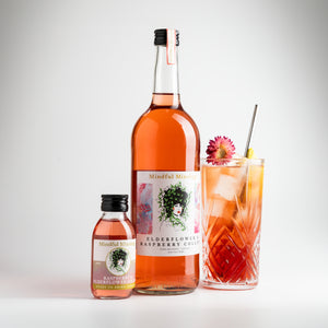 The small Raspberry and Elderflower gin Collins bottle is in the picture, alongside a large 750ml bottle. It is the same shape and similar label, but the bottle is a lot larger. Next to it is a glass of Raspberry and Elderflower gin Collins in a tall crystal glass. The cocktail is red in colour, there are ice cubes in the drink. It is garnished with a pink straw flower, a slice of lemon and a stainless steel straw. The background is white.