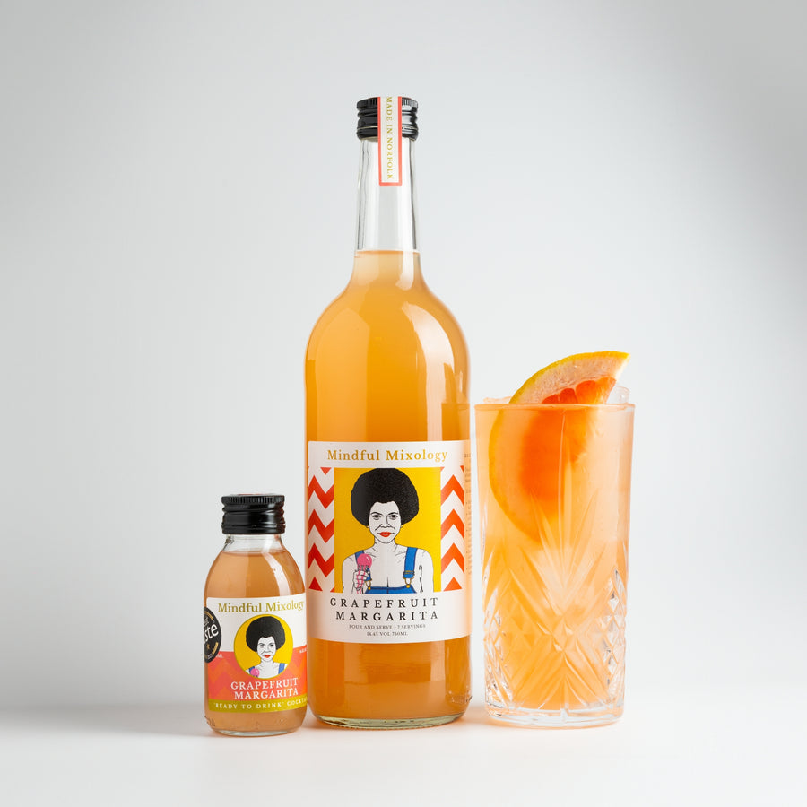  The small Grapefruit Margarita bottle is in the picture, alongside a large 750ml bottle. It is the same shape and similar label, but the bottle is a lot larger. Next to it is a glass of Grapefruit Margarita served as a Paloma. The glass is crystal and tall. The drink is orangey/pink, there is ice in the glass and it looks nicely chilled. There is a large grapefruit slice poking out of the top. The background is white.