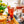 Load image into Gallery viewer, There is a picture of a raspberry and elderflower collins in a crystal tall glass, with lots of ice, a flower garnish, lemon wedge and a silver straw. There is a mini bottle of lychee martini on the table, a passio fruit martini and a lychee martini in someone&#39;s hand. There is a plant pot on the table with yellow flowers. The table is wooden and looks like it is outside. 
