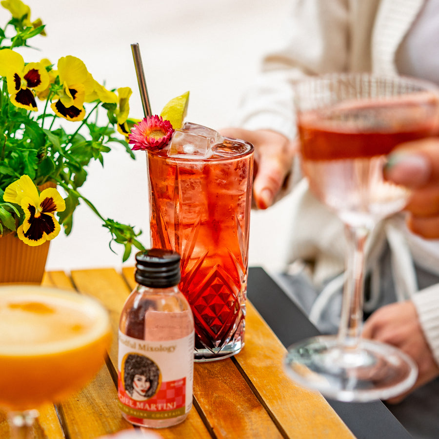 There is a picture of a raspberry and elderflower collins in a crystal tall glass, with lots of ice, a flower garnish, lemon wedge and a silver straw. There is a mini bottle of lychee martini on the table, a passio fruit martini and a lychee martini in someone's hand. There is a plant pot on the table with yellow flowers. The table is wooden and looks like it is outside. 