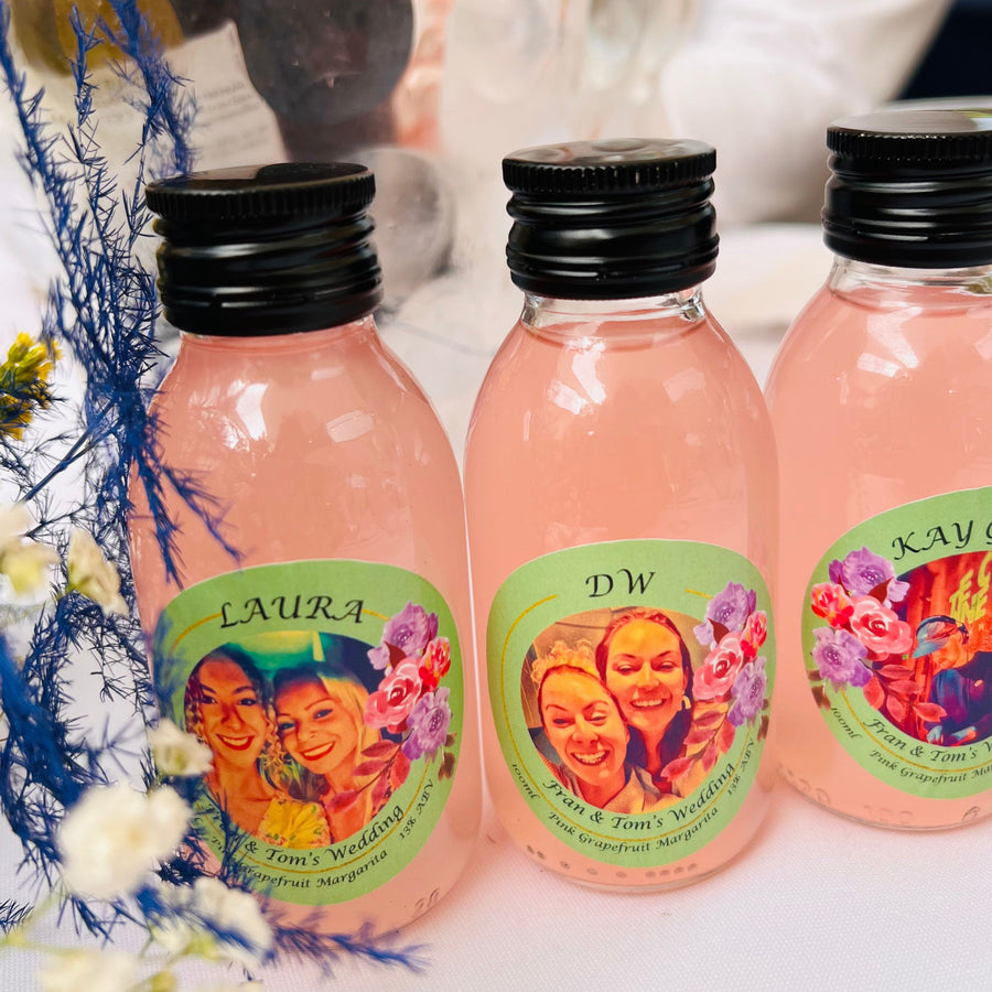 Picture shows three single-serve cocktails on a tablecloth. They are pink in colour. The bottles have a black cap. The label is green with flowers on and shows smiling faces. Each label has a different name, but all three cocktails are grapefruit margaritas. The header on each cocktail is the name of the person on the picture. 'Laura' is an example. Underneath it says 