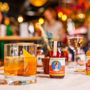 The Aperol Negroni single-serve, and the Aperol Negroni in a glass is shown on a table in what could be a house or a bar. There are two people in the background blurred out and a few nice lights in the background giving a warm hue. There are other drinks blurred out on the table, and what looks like a bowl of almonds. 