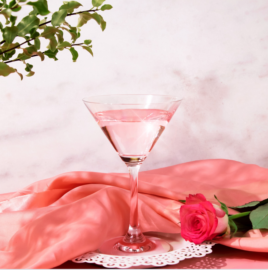 A lychee martini is shown in a martini glass. There is a grey background and a plant in the foreground. On the table is a pink tablecloth and a pink rose.